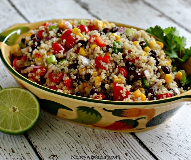 Shot of the quinoa salad prepared and ready to go in a festive dish with peppers on the side of it and limes cut in half next to the bowl.