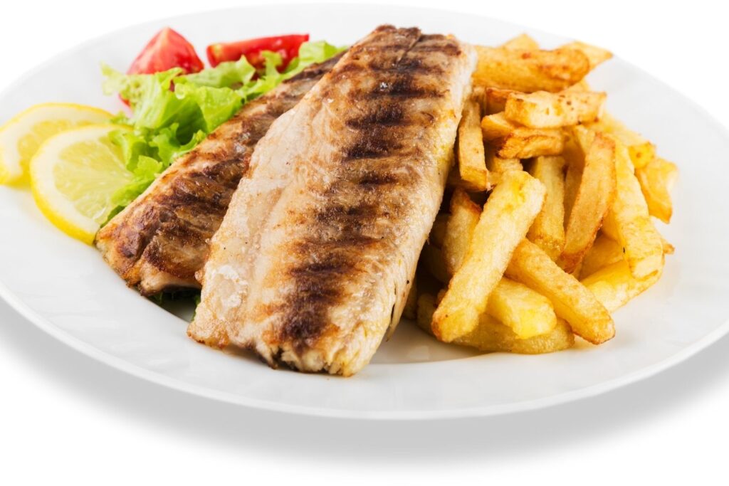 Grilled tilapia fillets on white plate with green salad lemon slices and french fries
